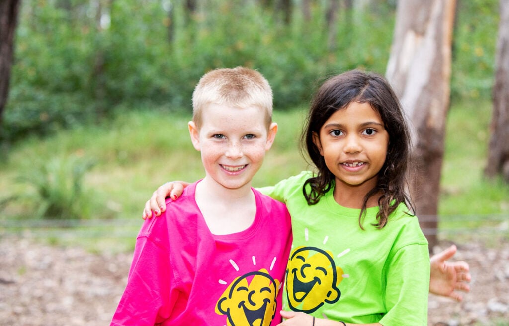 Two kids at a Camp Quality camp stand smiling with their arms around each others shoulders