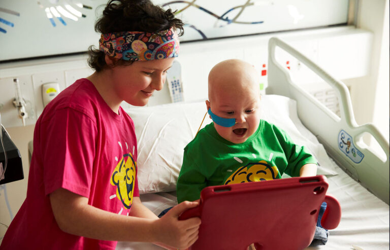 Two Camp Quality kids sat on a hospital bed using a tablet device