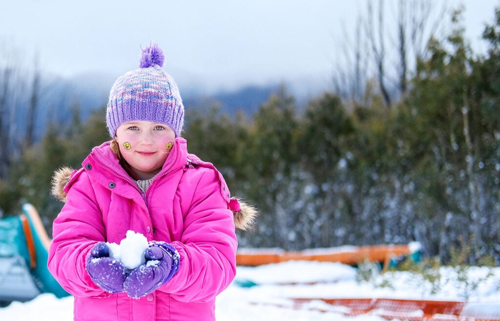 A child is wrapped up warm in pink snow gear, holding a snowball