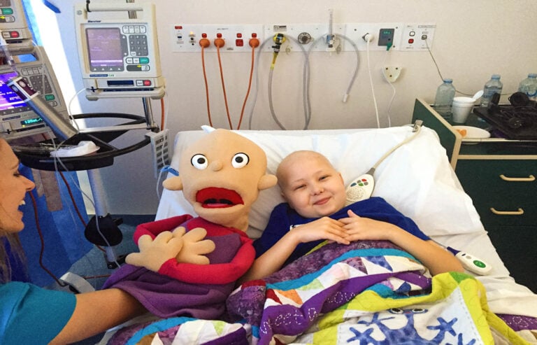 A child in a hospital bed with a Camp Quality puppet next to them