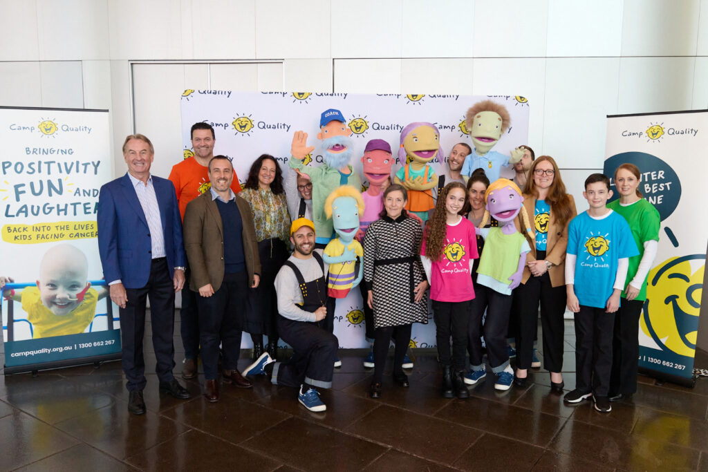 A bunch of people stand and smile in front of a media wall with a puppet