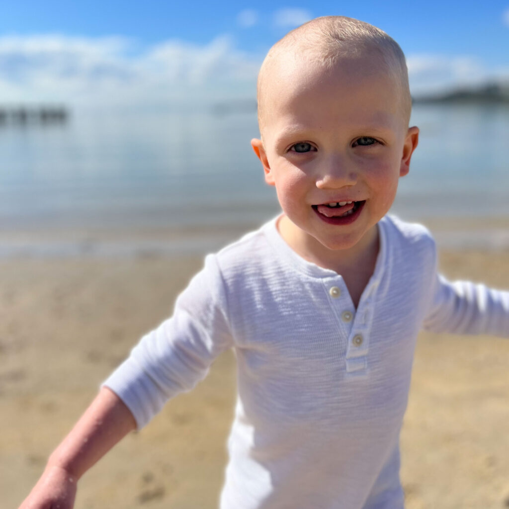 Toddler with shaved head smiles on a sunny beach