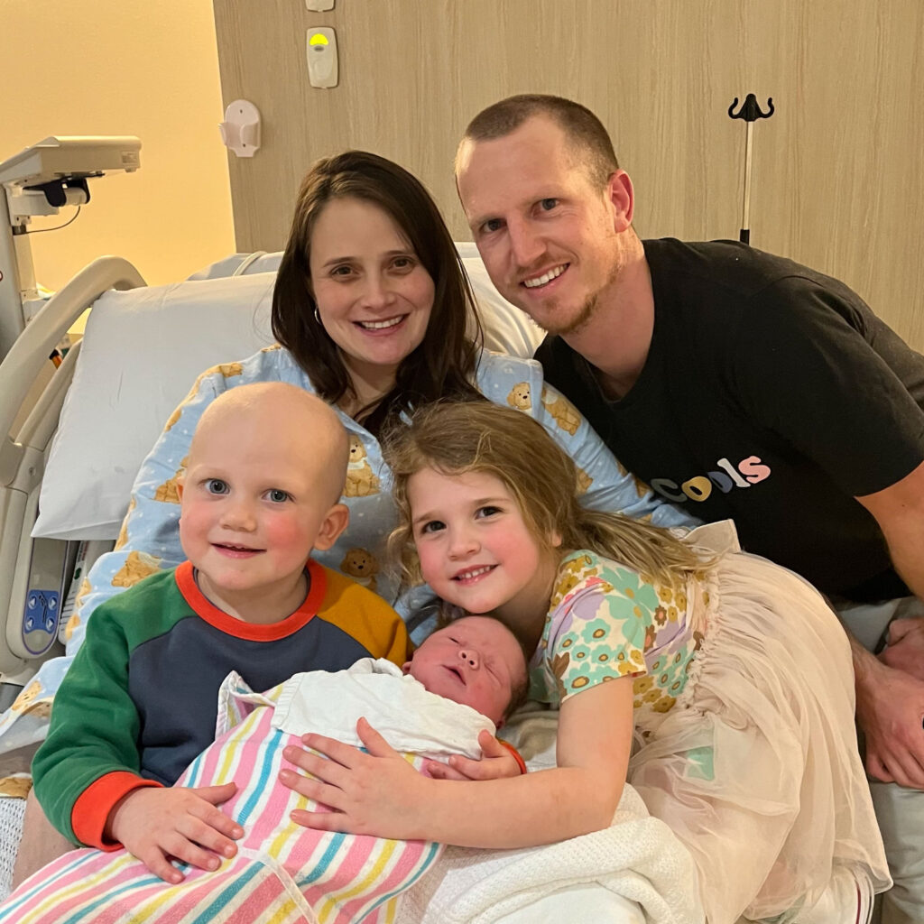 A family smiles gathered around the mum in a hospital bed and a newborn baby. One boy has a shaved head.