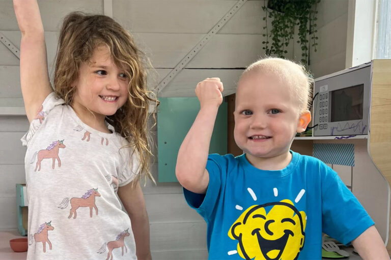 Two young kids, one with a shaved head smile with arms in the air