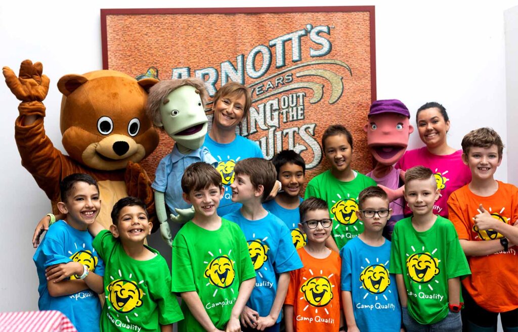 Children smiling together in Camp Quality shirts in front of an Arnotts sign