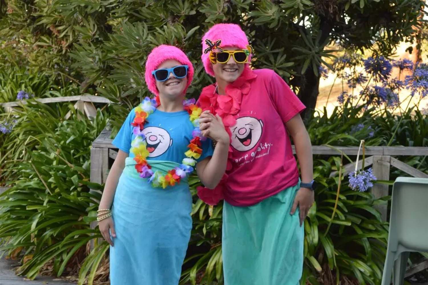 Two girls in matching pink wigs and mermaid tails towels smile